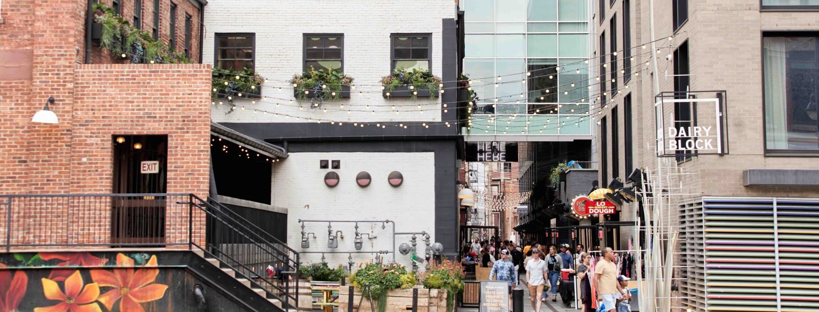 Urban alley featuring string lights, brick buildings, and people walking near a "Dairy Block" sign