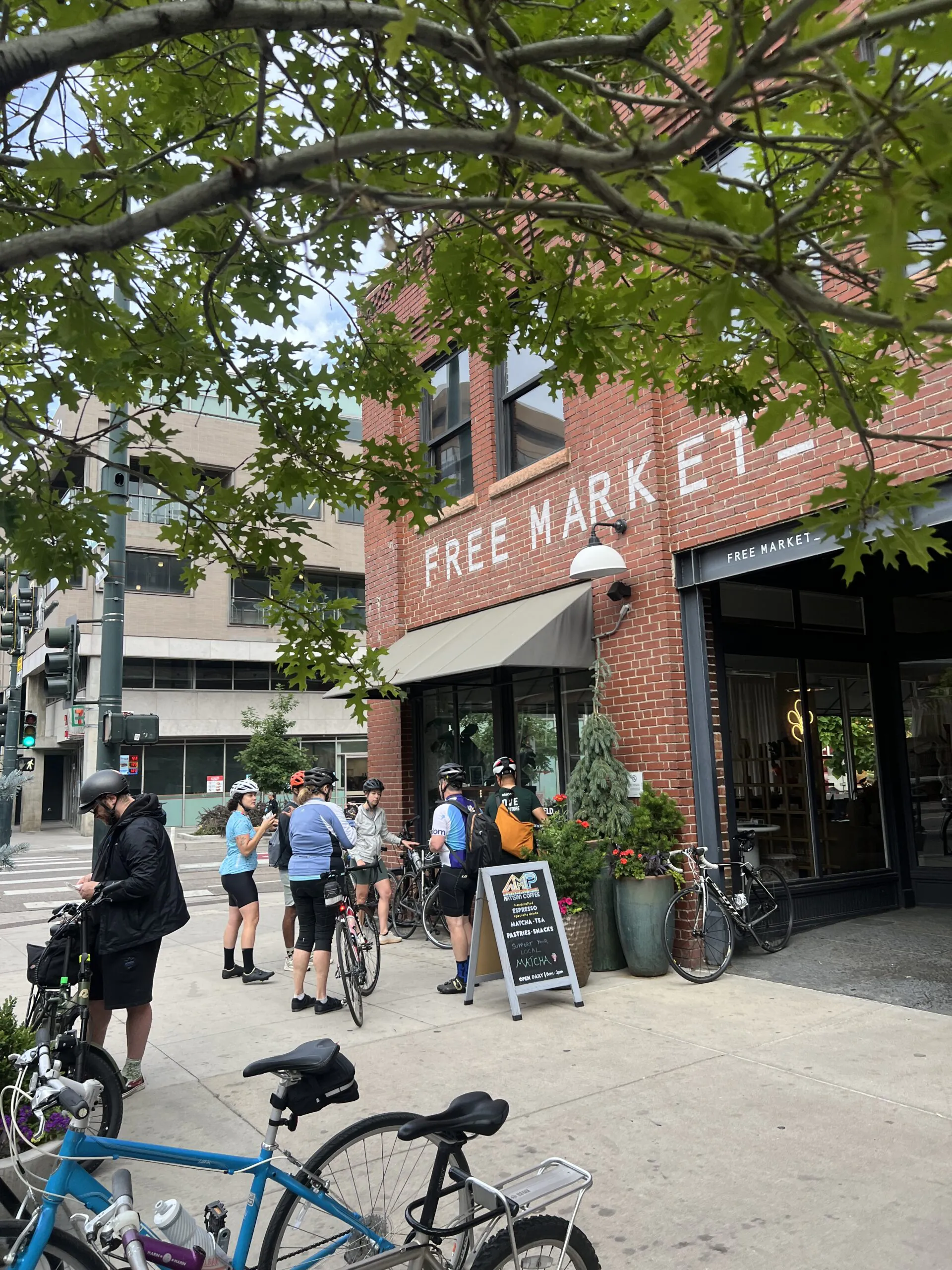 Cyclists gather outside "Free Market" brick building under leafy tree branches