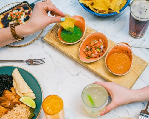 Tacos, margaritas, and salsas on table with hands grabbing food