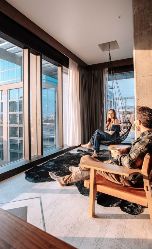 Man and woman sitting in Maven hotel room looking out large glass windows