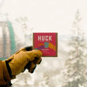 Gloved hand holding HUCK coffee pack, snowy mountain background