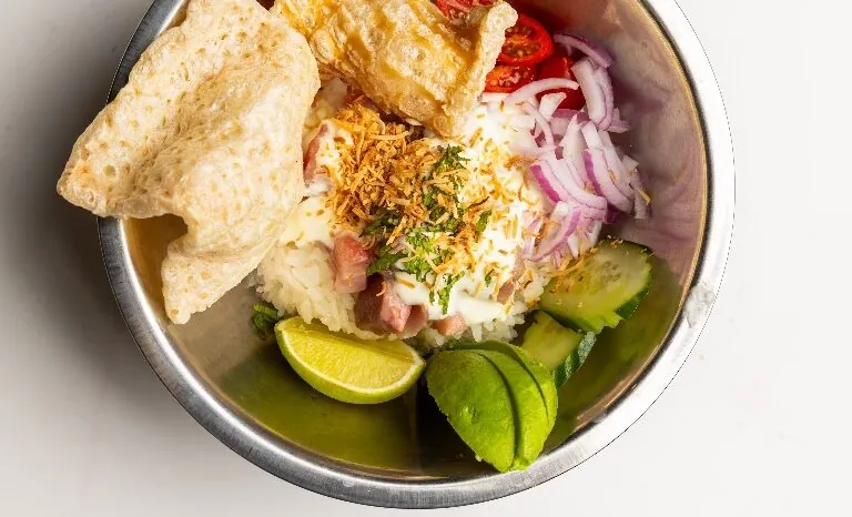 Bowl of rice topped with sliced onions, cucumber, lime, red chili, and crispy pork rinds