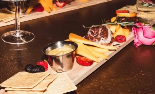 Charcuterie board with cheese, meats, crackers, strawberries, black olives, mustard, and a pink rose garnish on a wooden table