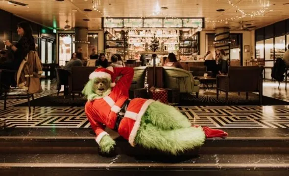 Person in Grinch costume lounging inside festive restaurant, surrounded by people dining