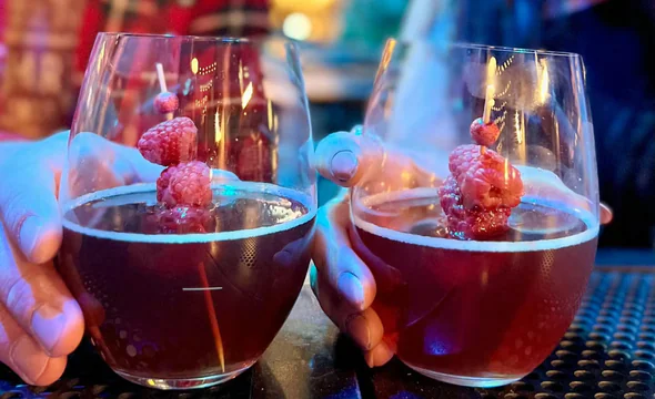 Two glasses of red cocktails garnished with skewered raspberries, held by two people at a bar