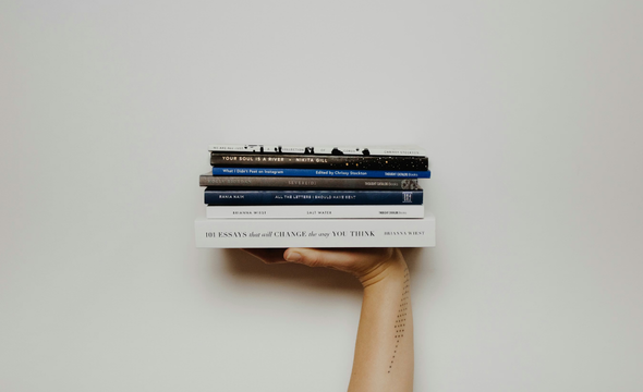A tattooed arm extends to hold a stack of five books, including 
