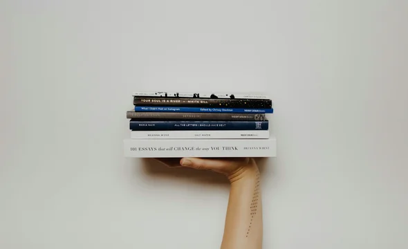 A tattooed arm extends to hold a stack of five books, including 