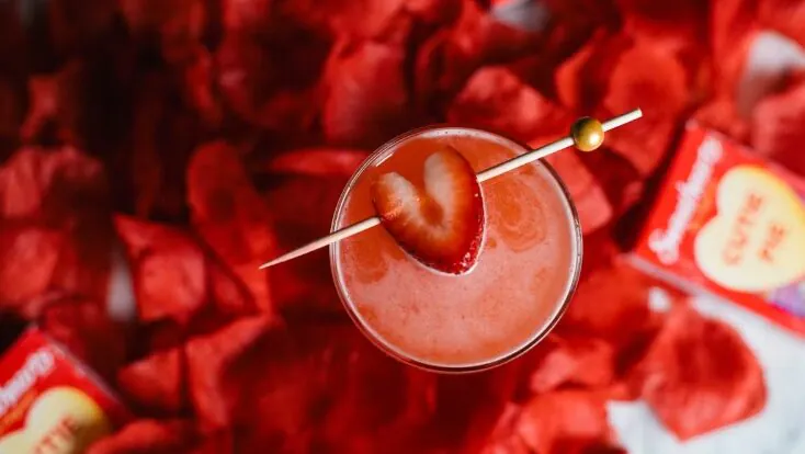 Heart-shaped strawberry garnish cocktail surrounded by red rose petals, with 